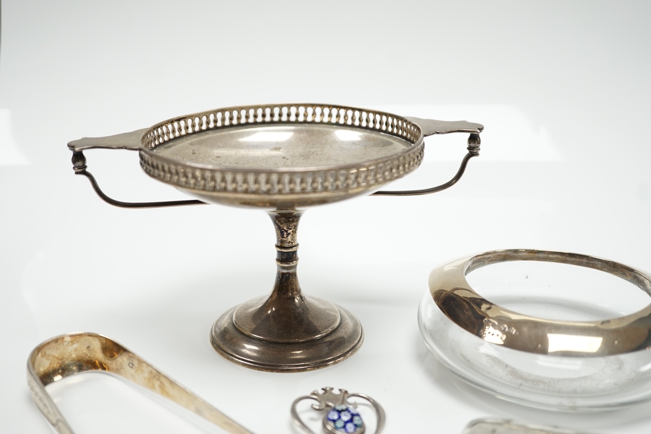 A George V silver tazza, a silver cigarette case, a pair of George III silver sugar tongs, a silver mounted glass shallow dish and a modern silver and lass inset pendant.